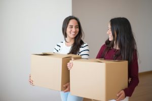 two women smiling at each other while carrying moving boxes